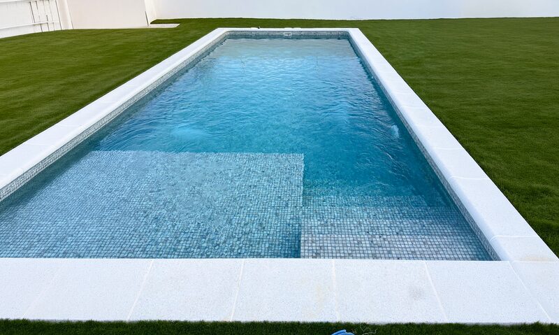 Piscina rectangular con gresite azul claro, banco interior y césped alrededor